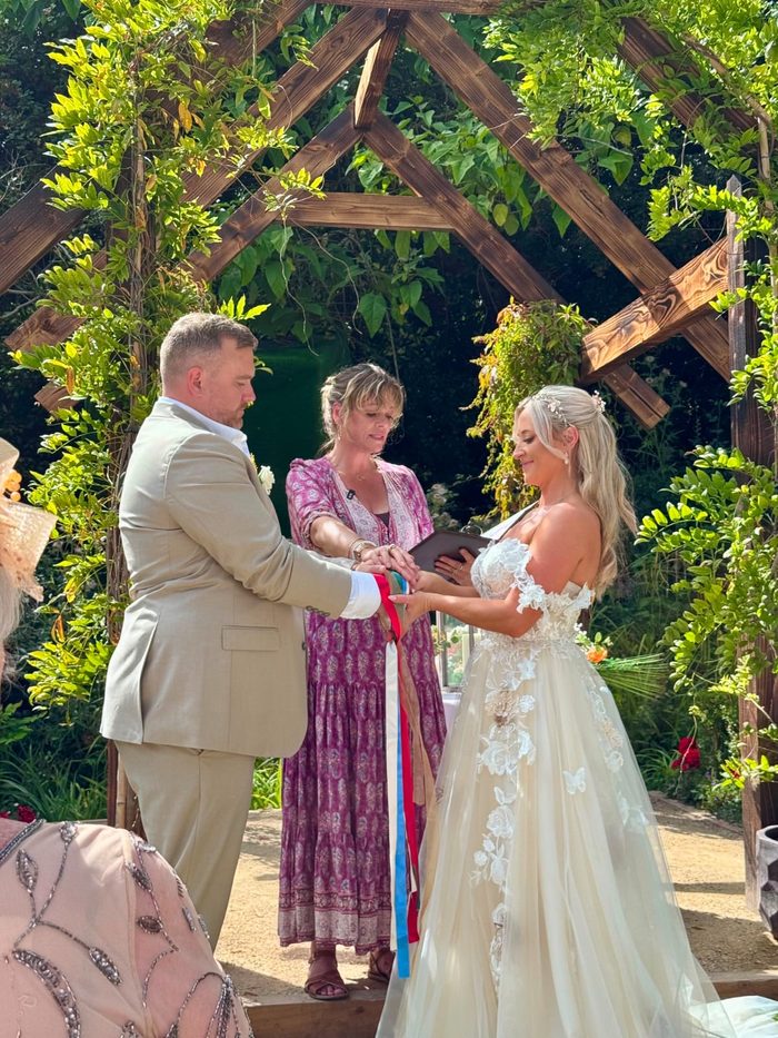 Emma holding her ceremony folder as a couple exchange vows beneath a wooden garden arbour, their hands bound with ribbons.