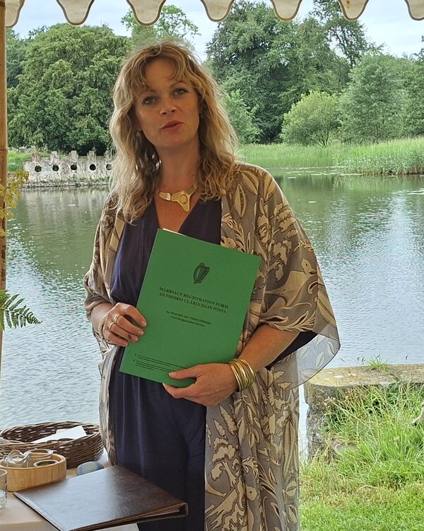 Emma O'Brien beside a lake under an open marquee, holding the green Irish marriage registration form.