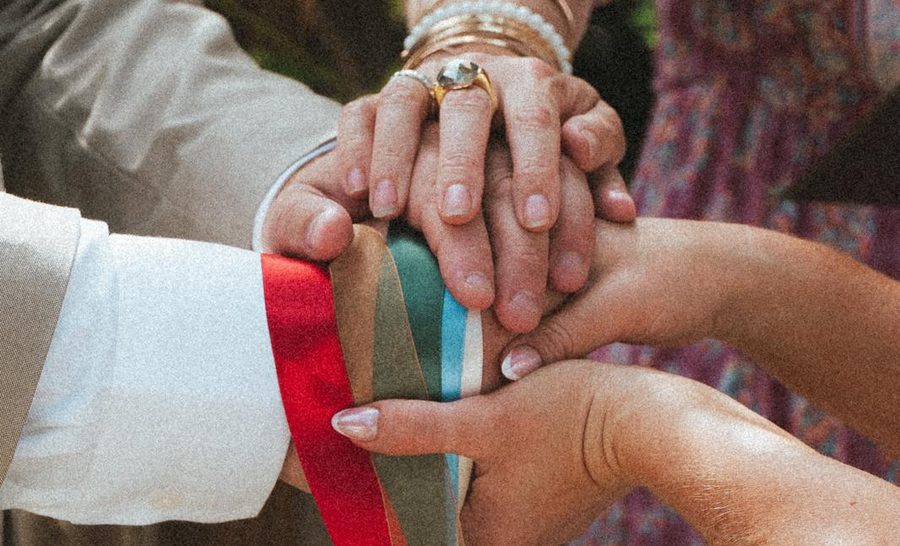 Emma's hands resting over a couple's joined hands, bound together with coloured handfasting ribbons.