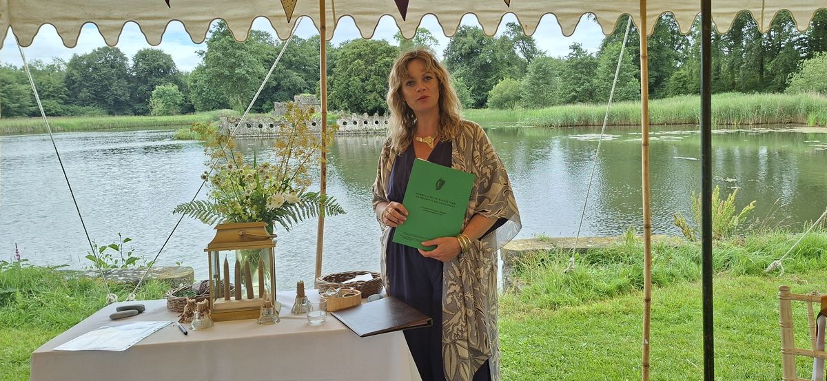 Emma O'Brien standing beside a lake under an open marquee, holding the marriage registration form, with candles and wildflowers on the table beside her.
