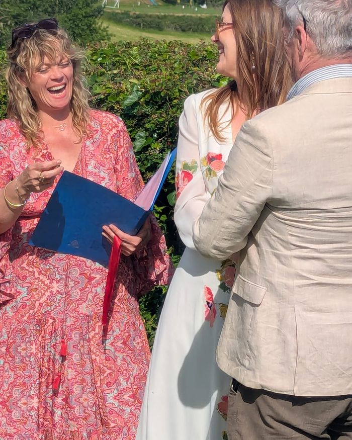 Emma laughing with her head thrown back while conducting a ceremony for a couple in a garden.