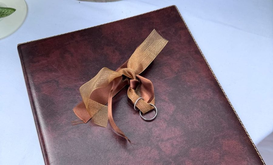 A burgundy leather ceremony folder on a linen table, tied with a gold ribbon with a wedding ring threaded through it.