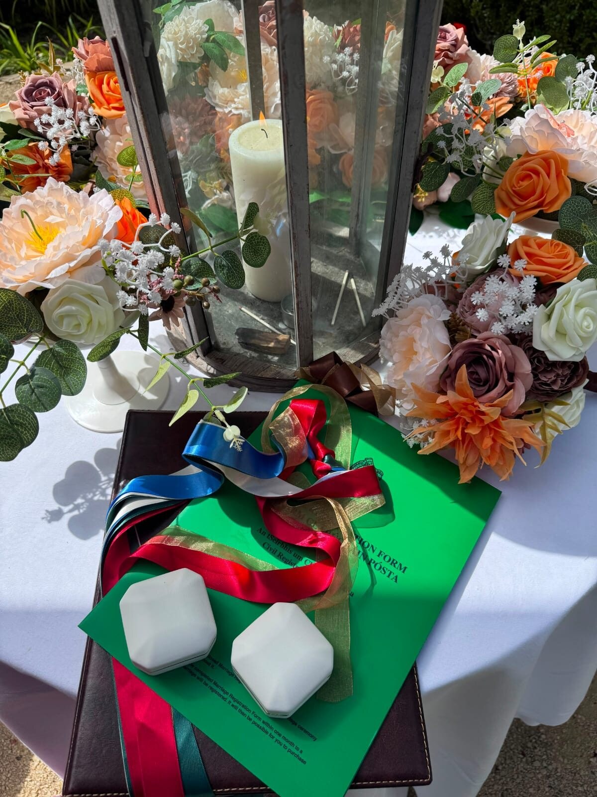 A ceremony table set for a wedding: the couple's ring boxes and coloured handfasting ribbons resting on the green Irish marriage registration form, beside a candle lantern and summer flowers.