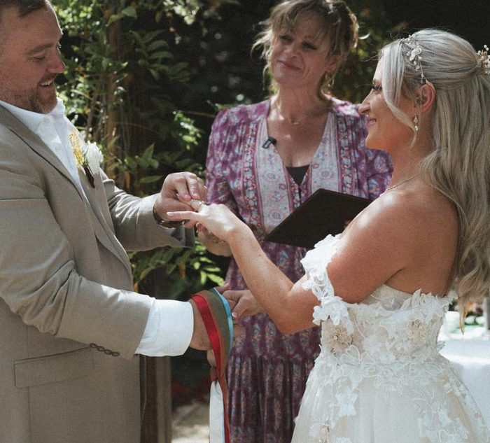 Emma looking on as a groom slides a ring onto his bride's finger.